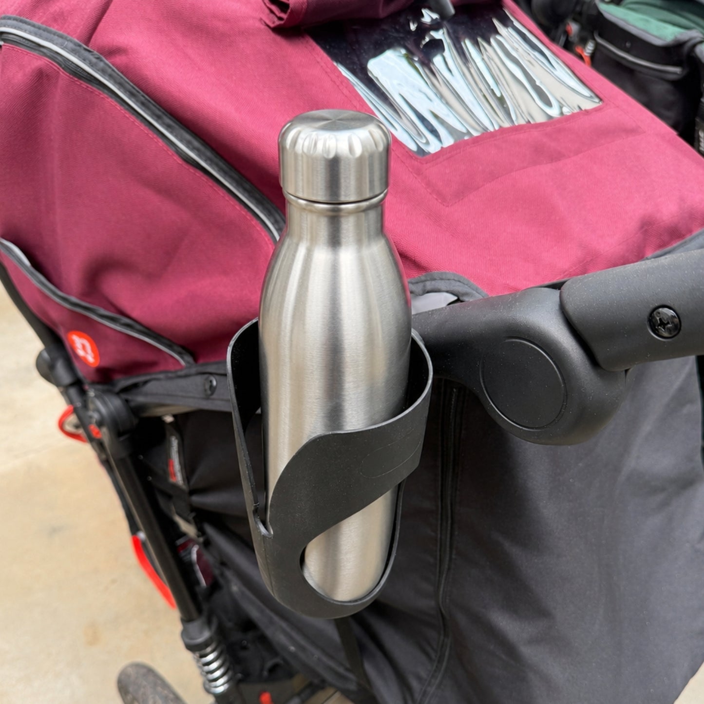 A stainless steel water bottle held in a black cup holder attached to an Out 'n' About Nipper stroller with a burgundy fabric canopy.