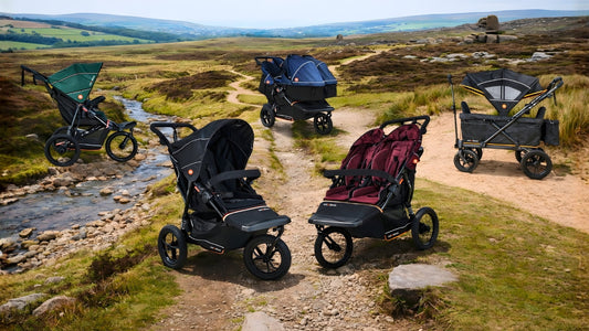 A group of out ‘n’ about all-terrain buggies and pushchairs displayed on a rugged countryside path beside a shallow stream, with rolling hills, rocky moorland, and open rural scenery in the background