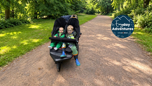 A Summit Black Out 'n' About Nipper Double V6 pushchair with baby and a toddler seated inside, on a gravel path in a park. A blue circular 'Everyday Adventures' logo is visible in the top right corner.