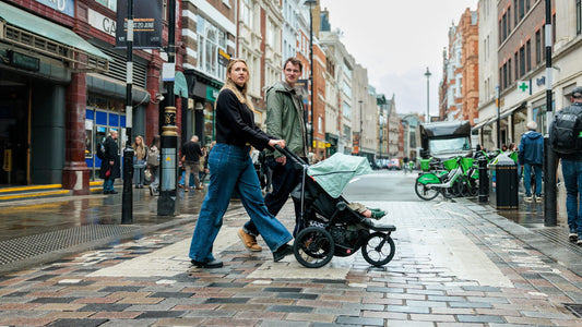 A woman pushing the Pebble Mint Nipper Compact buggy with a toddler, while a man walks beside her across a busy cobbled city street in London