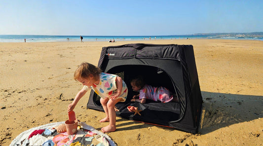 Toddlers playing on a beach beside an Out’n’About Nipper Nest travel cot