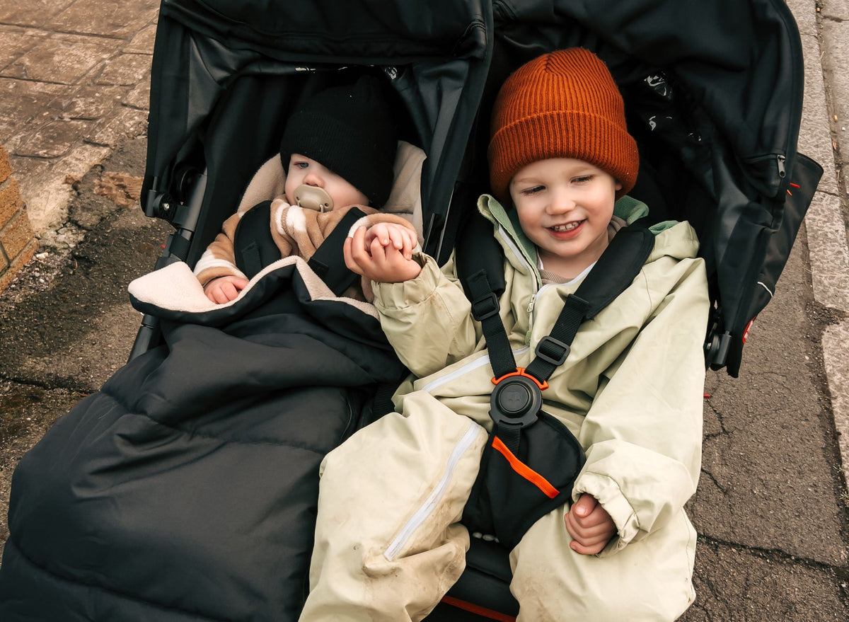 Two children in cream footmuffs looking up from the seats of a Nipper Double pushchair