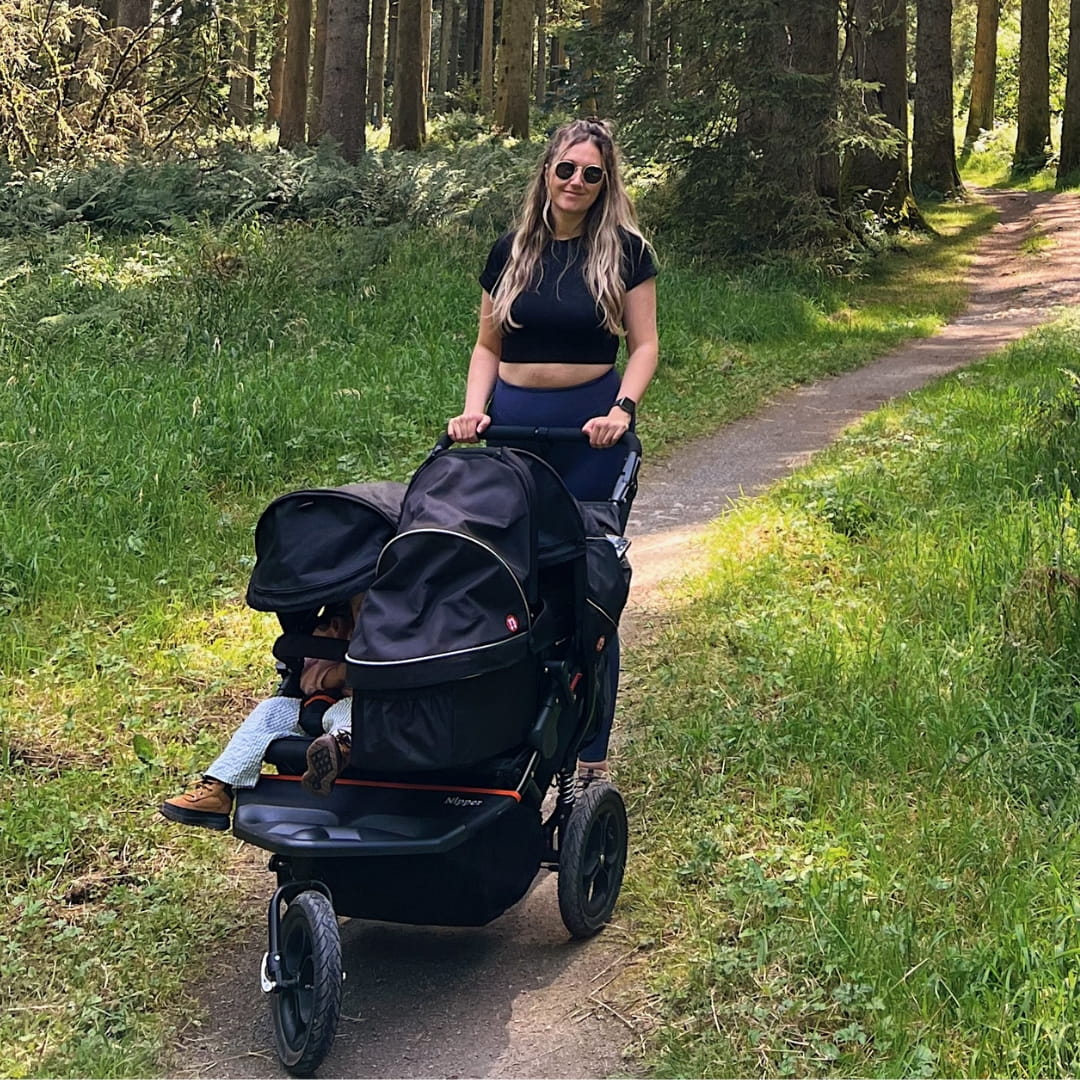 Parent pushing a Nipper Double V5 double stroller for newborn and toddler along a woodland trail